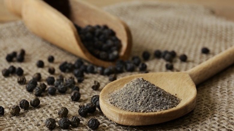 Ground black pepper on a wooden spoon surrounded by peppercorns.