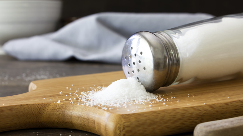 A saltshaker on its side with a pile of salt on a cutting board