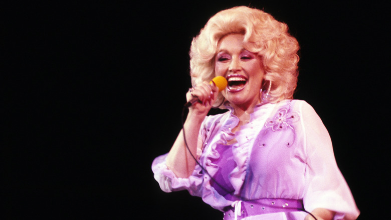 Dolly Parton in a purple dress holding a microphone and singing.