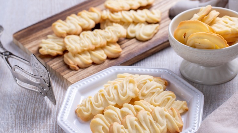 Freshly baked homemade spritz cookies set out on a plate, in a bowl, and on a cutting board next to tongs.