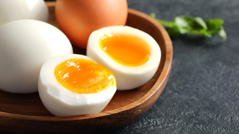 A halved soft-boiled egg next to whole peeled and unpeeled eggs on a rimmed wooden plate