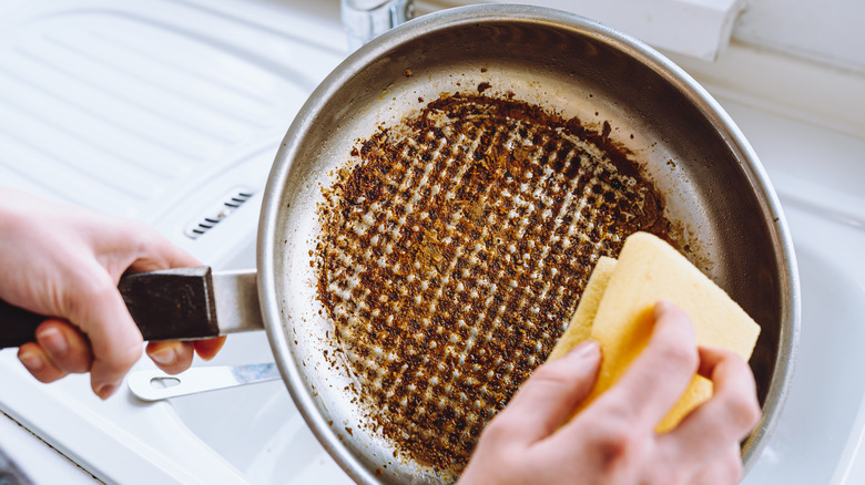 A person cleaning a pan with scorch marks and food debris using a sponge