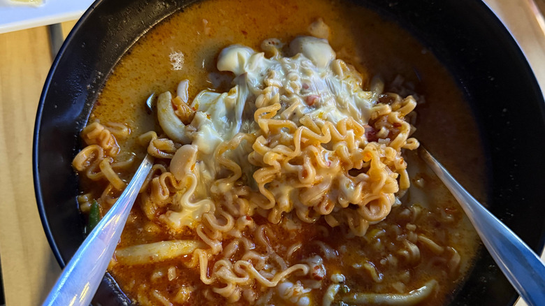 Black bowl of spicy soup and creamy noodles with added cheese and two spoons.