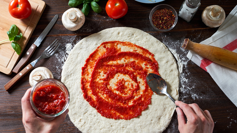 Hands spreading sauce on pizza crust with toppings surrounding it