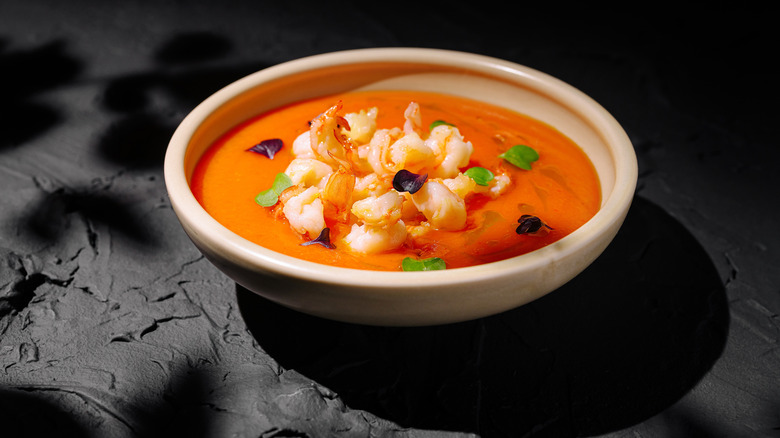 A bright orange and red seafood bisque topped with sautéed shrimp and herbs on a black stone countertop.