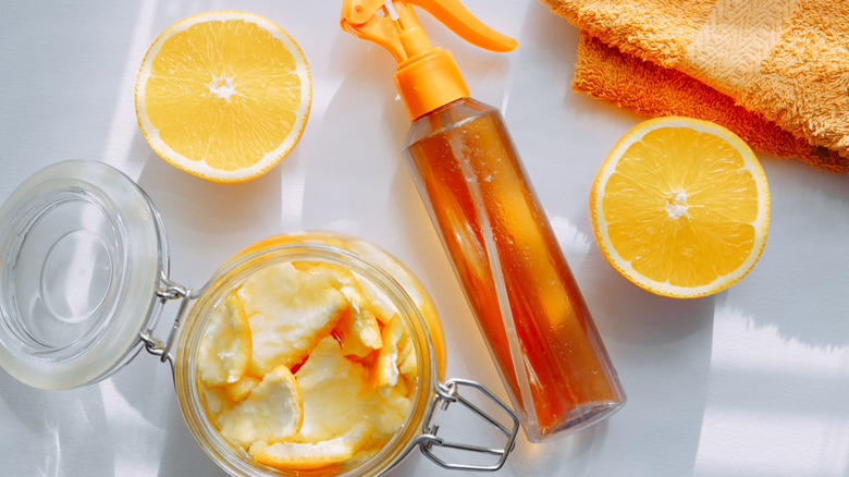 Orange peels in a jar, a spray bottle full of a clear brownish liquid, and an orange cut in half.