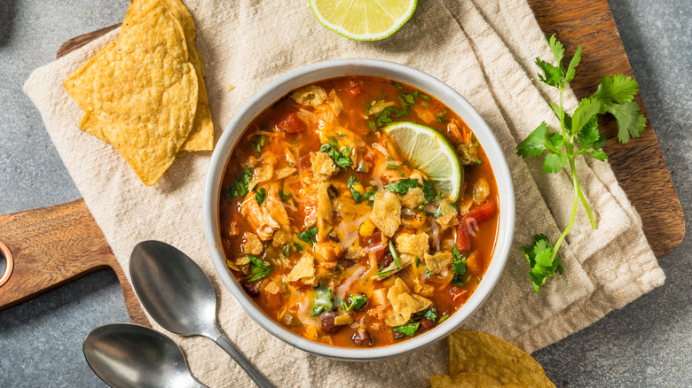 Chicken tortilla soup in a white bowl on a cutting board with cilantro, a lime, and chips