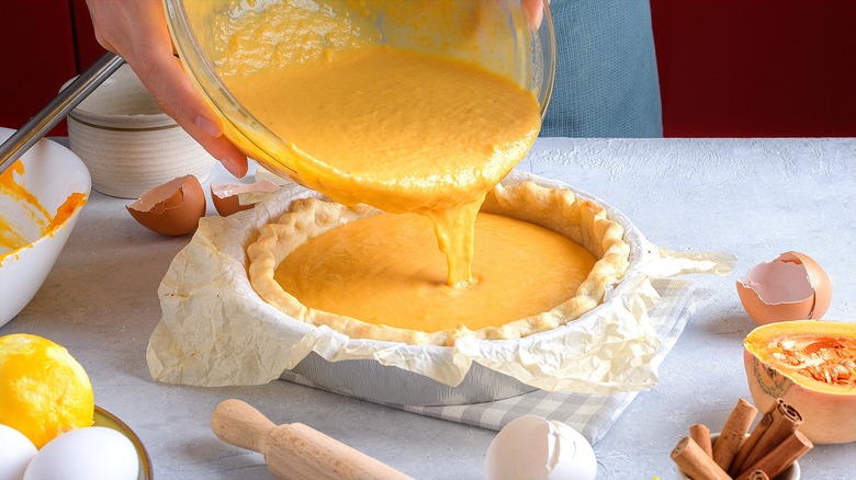 Hands pouring a bowl of creamy butternut squash pie filling into a par-baked pie crust
