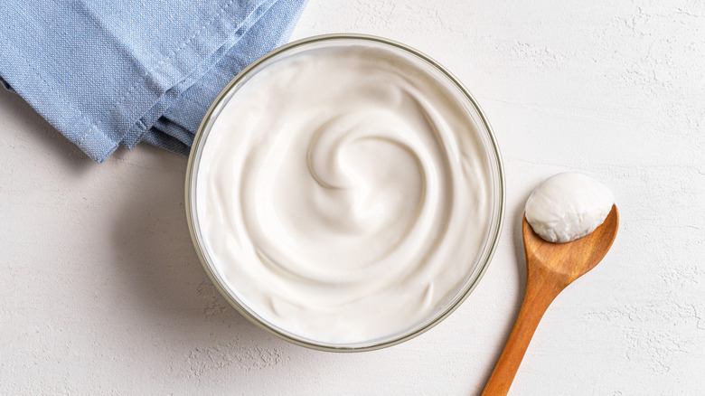 A bowl of plain yogurt with a wooden spoon and a blue linen napkin on a white surface.