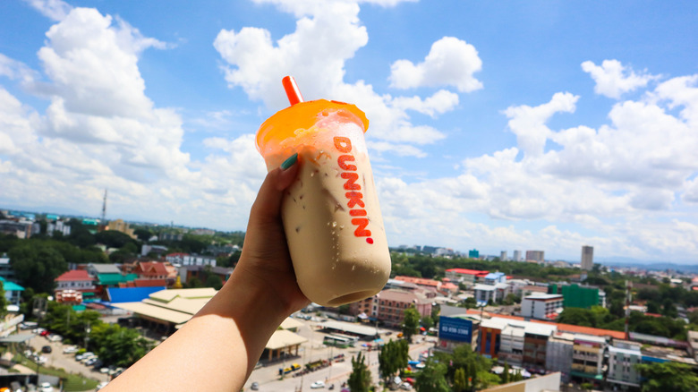 Woman's hand holding Dunkin' iced coffee in front of background of blue sky and city.