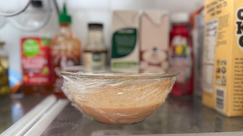 Dynamite sauce in glass bowl covered with plastic wrap in refrigerator