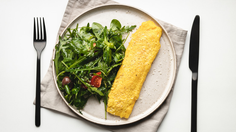 An omelet is served with greens on a white plate with a cloth napkin, fork, and knife.