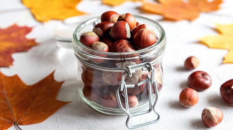 Acorns in a small glass jar surrounded by fall leaves on a white surface