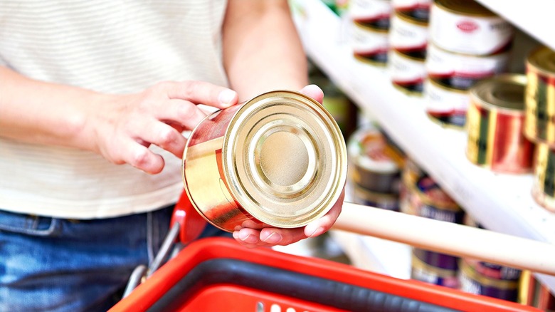 Two hands holding a gold can of food over a red shopping cart