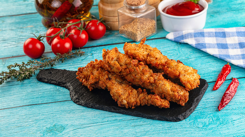Fried chicken strips on a black slate cutting board on a blue picnic table with tomatoes, herbs, and chili peppers.