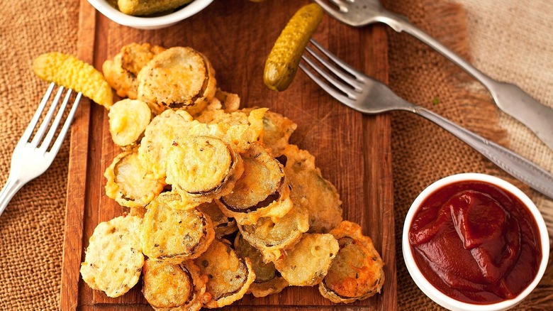 Fried pickle rounds on a wooden serving board near a small bowl of ketchup and cornichons on metal forks