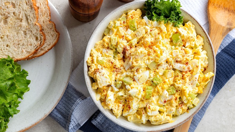 Egg salad with paprika and salad greens in a white bowl next to a plate with two slices of whole-grain bread.