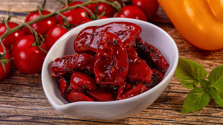 Sun-dried tomatoes in a small white bowl on a wooden table with basil leaves and fresh tomatoes in the background