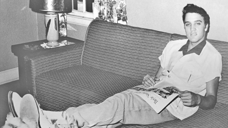 Young Elvis Presley reading a newspaper on a couch.