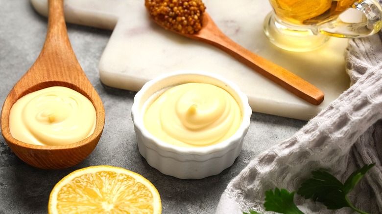 Dijonnaise in a small white bowl and on a wooden spoon next to half of a lemon and a spoonful of whole-grain mustard