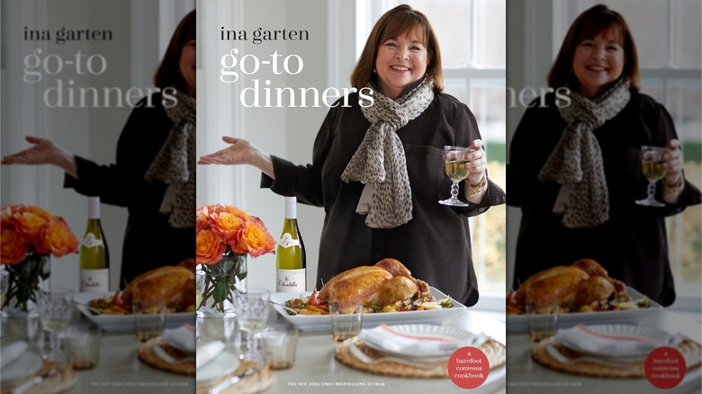Book cover for Go-To Dinners, showing Ina Garten holding a glass of wine while standing behind a table with a roast chicken on a white tray and a bottle of wine