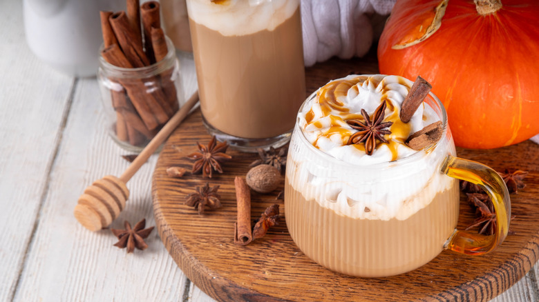 A pumpkin spices latte drink in a glass mug on a wooden table with various props behind it out of focus including a pumpkin and some cloves and star of anise