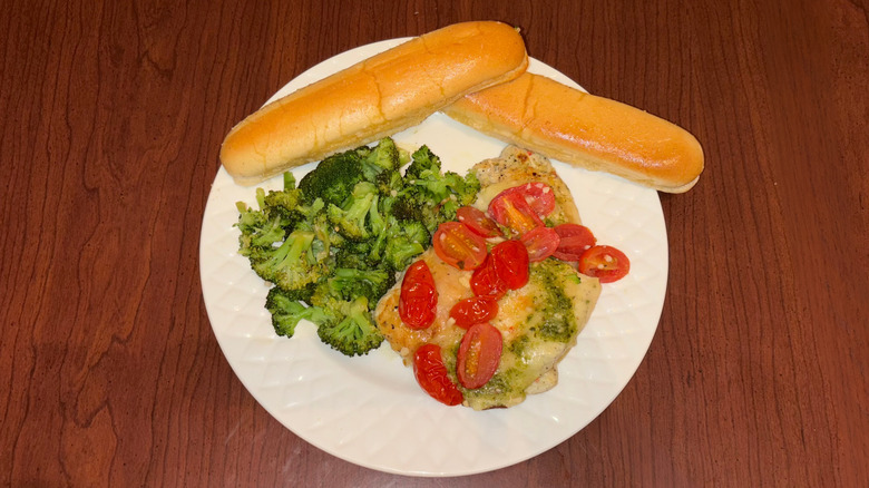 Chicken with cherry tomatoes and pesto on a white plate with broccoli and breadsticks.