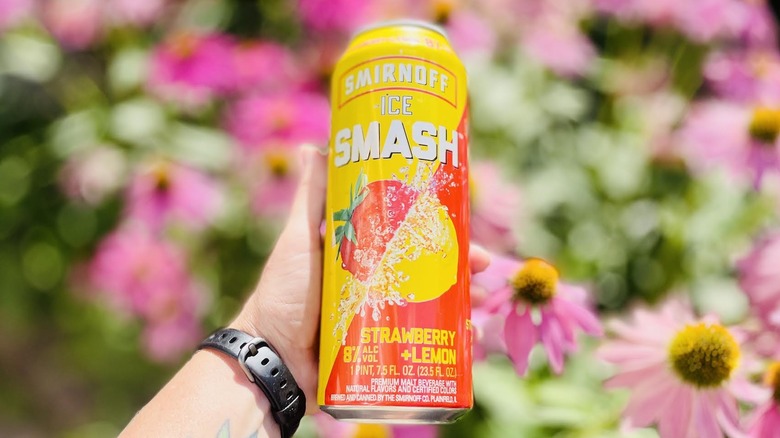 Hand holding can of Smirnoff Ice Smash Strawberry Lemon in front of blurred flowers