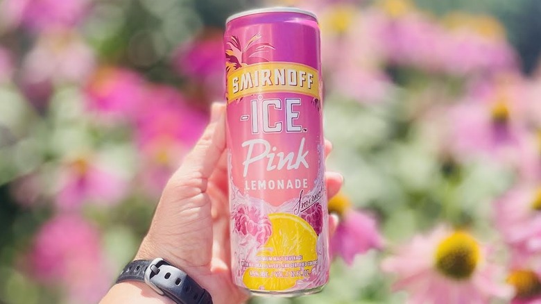 Hand holding can of Smirnoff Ice Pink Lemonade in front of blurred flowers