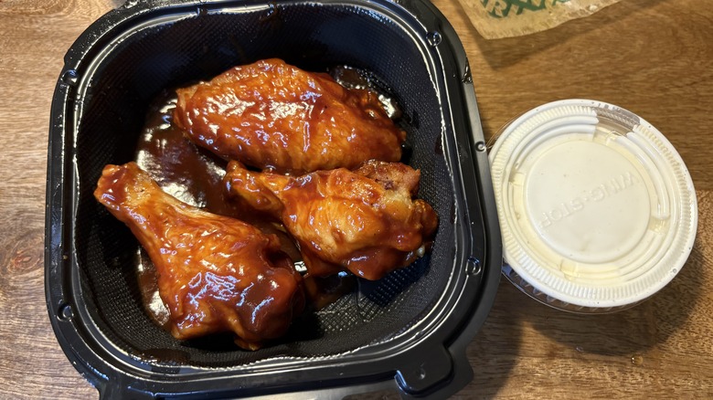 Three Hickory Smoked BBQ wings in a black to-go container beside a sauce cup