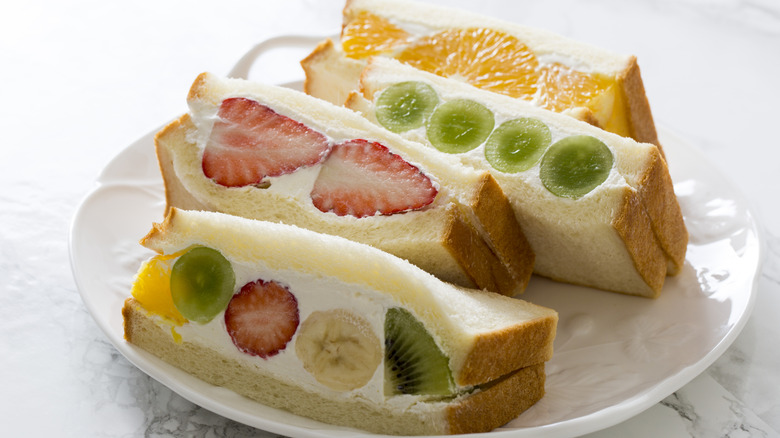 A selection of fruit sandwiches sliced into rectangles with strawberries, bananas, grapes, oranges, and kiwi on a white plate