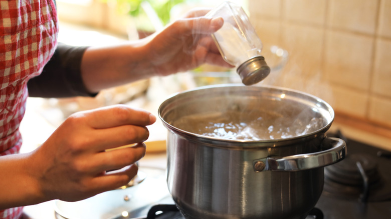 A cook holds a salt shaker over a pot of boiling water on the stove.