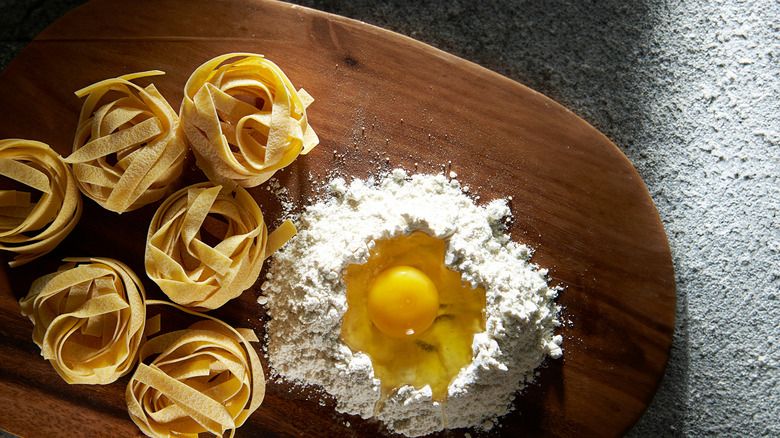 An egg yolk sits on top of a mound of white flour, with fresh pasta noodles nearby.