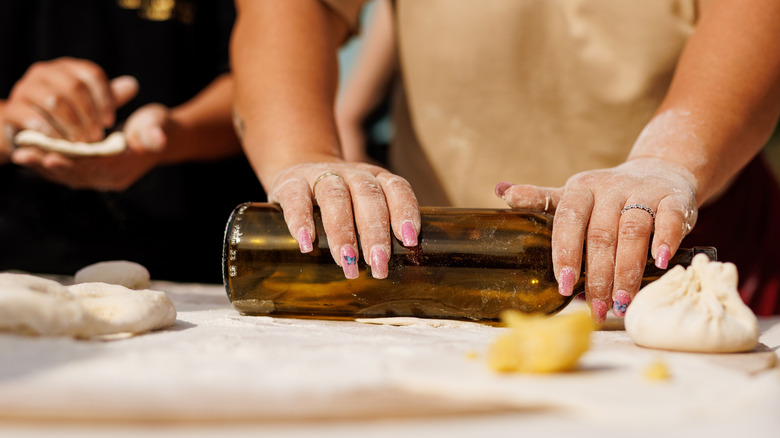 A woman's hands with long nails uses a wine bottle as a rolling pin.