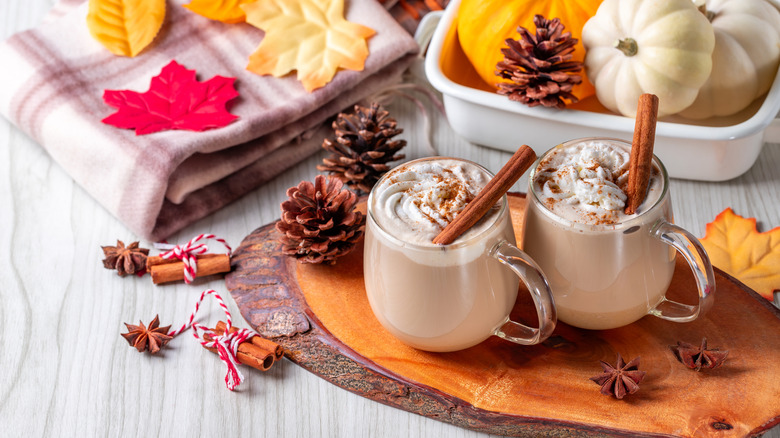 Two pumpkin spice lattes surrounded by whole pumpkins, leaves, and spices