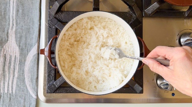 Fluffing rice grains with fork in brown pot on stovetop