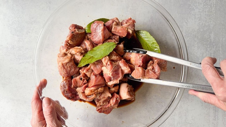 Tossing pork shoulder cubes with adobo marinade using tongs in large glass bowl