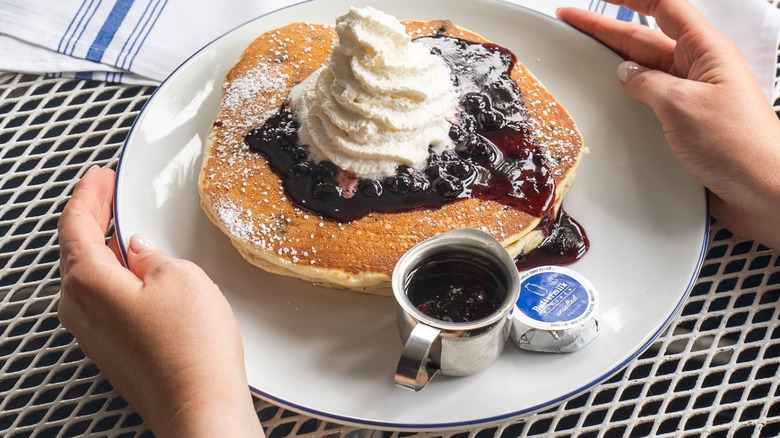 Hands holding plate of blueberry pancakes topped with whipped cream