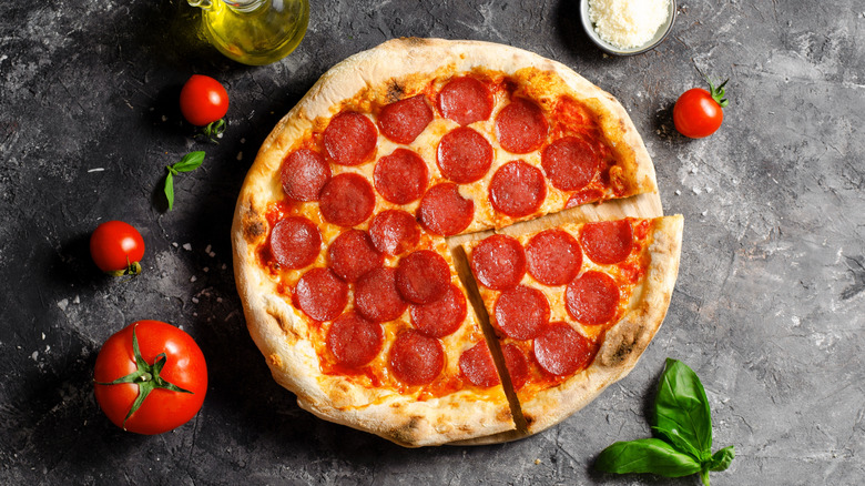 A pepperoni pizza is arranged on a rustic table eith tomatoes, basil leaves, olive oil and Parm.