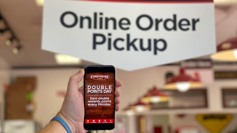 Hand holding phone with Firehouse Subs' app on screen in front of a to go sign for online order pickup