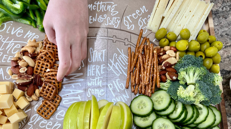 Person preparing a football-themed charcuterie board with veggies, fruits, nuts, pretzels, olives, and cheese