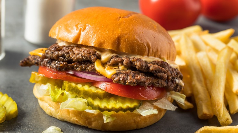 A smash burger with multiple patties, onions, pickles, tomatoes, and cheese with some fries next to it.