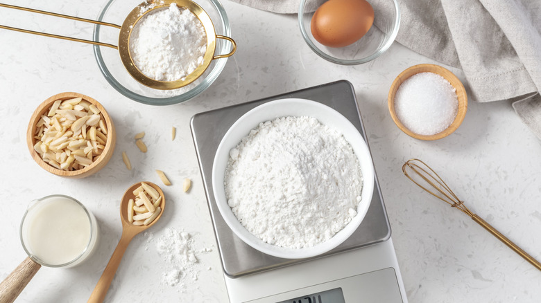 A variety of baking ingredients separated on a countertop with a bowl of flour on a digital scale