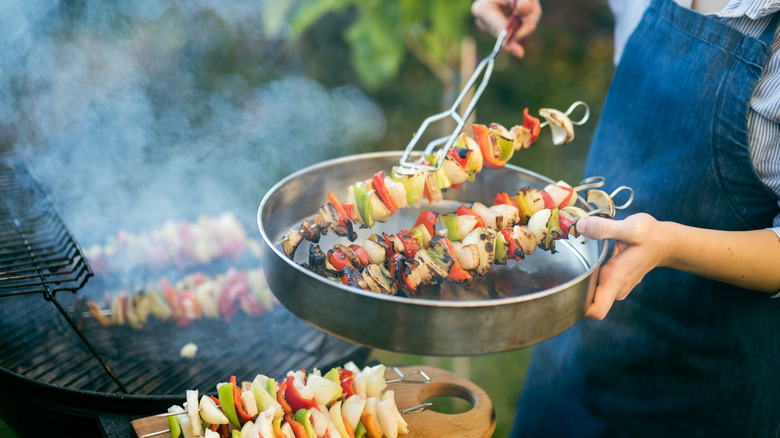 A person holding a metal bowl of skewers, about to transfer them to the grill with tongs