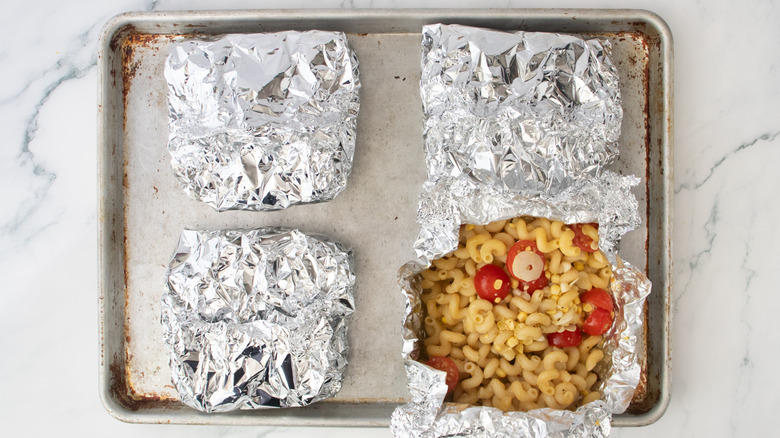 Four grilled parcels of pasta in aluminum foil on a baking sheet, one is opened