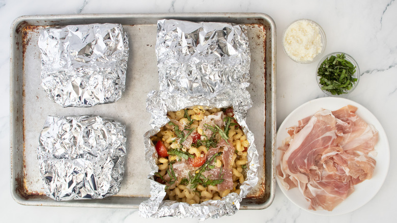 Four grilled parcels of pasta in aluminum foil on a baking sheet, one is opened and topped.