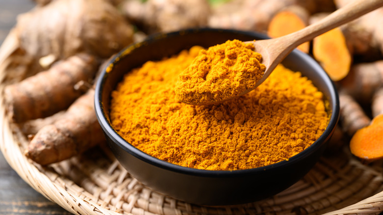 Ground turmeric in a bowl with a wooden spoon with sliced and whole turmeric roots in the background