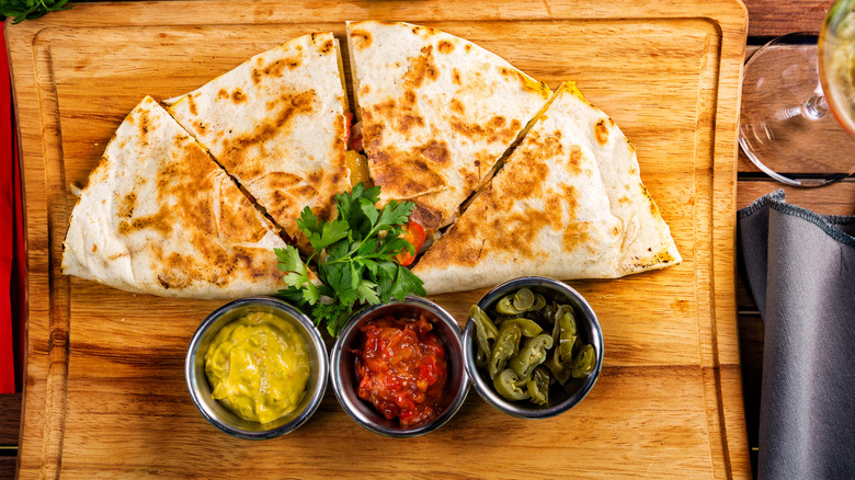 Quesadilla with cilantro, dipping sauce, salsa, and sliced peppers on a wooden cutting board
