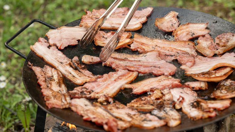 strips of bacon cooking in a cast iron pan over an open fire