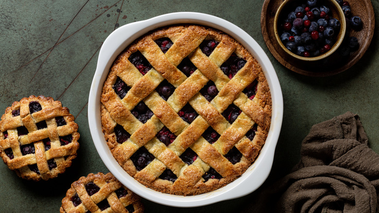 Blueberry pie in a dish with two smaller pies and a bowl of frozen blueberries around